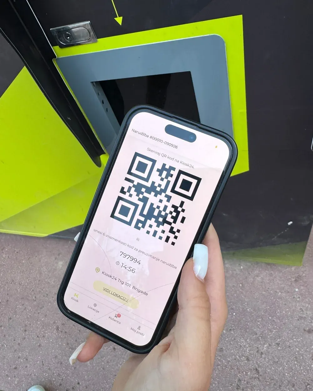 A hand holding a phone that shows a QR code, in front of a yellow and black Kiosk24 kiosk.